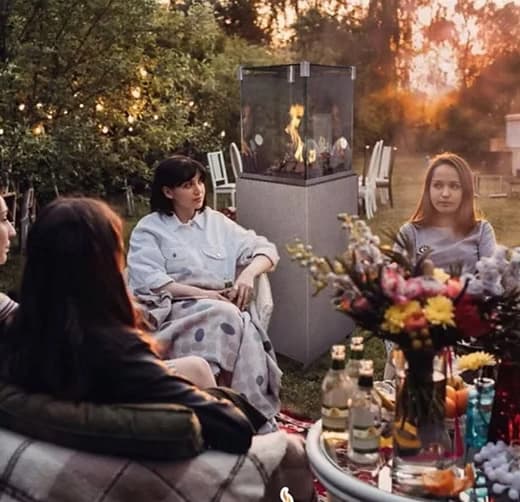 People seated around a glass-sided outdoor gas heater in a garden at dusk