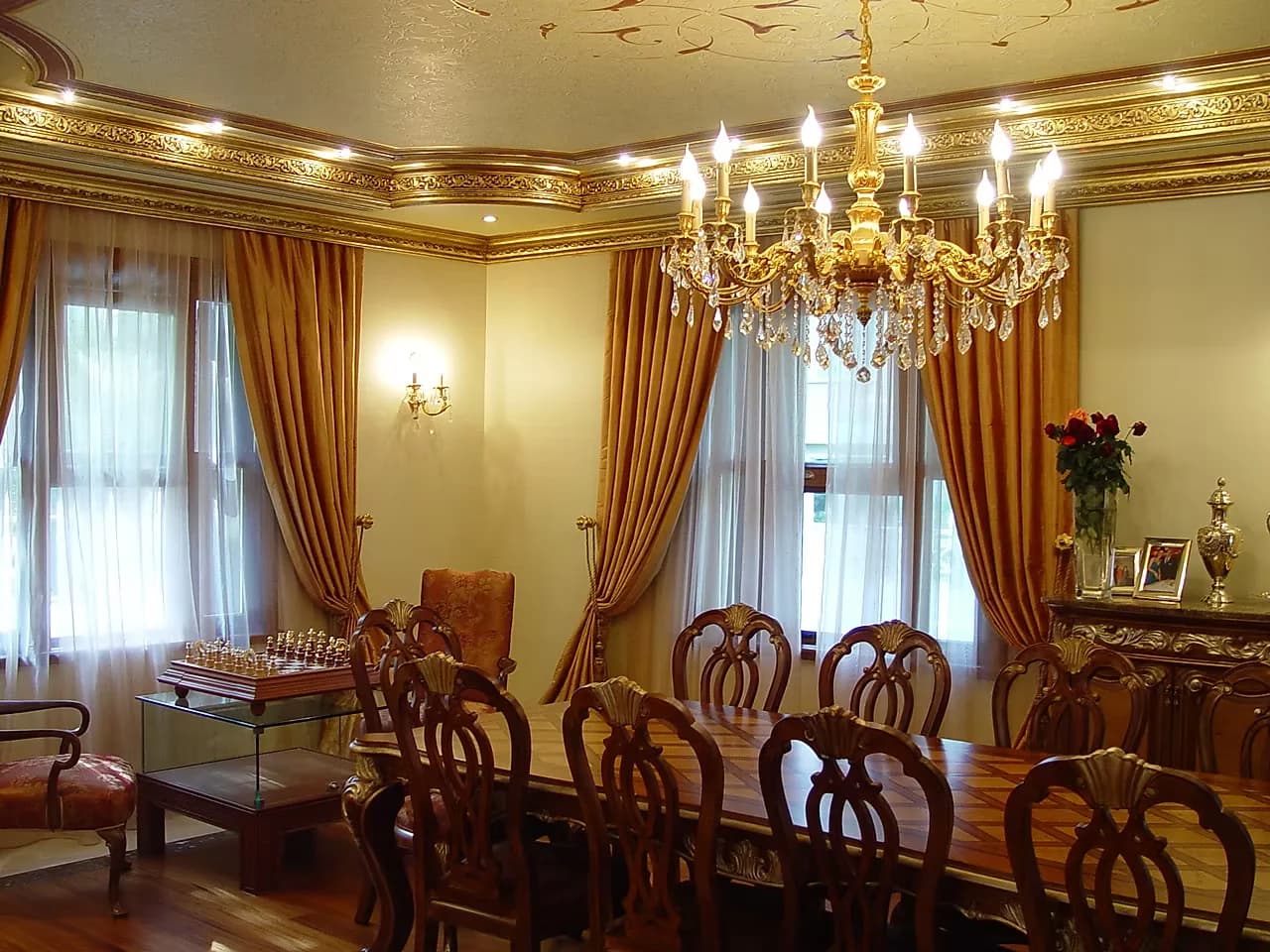 Dining room with a gilt chandelier, moulded cornice and a long carved table beneath gathered drapes