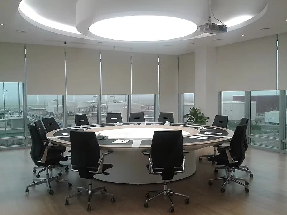 Circular board room with a ring table beneath a lit ceiling disc, glazed on three sides