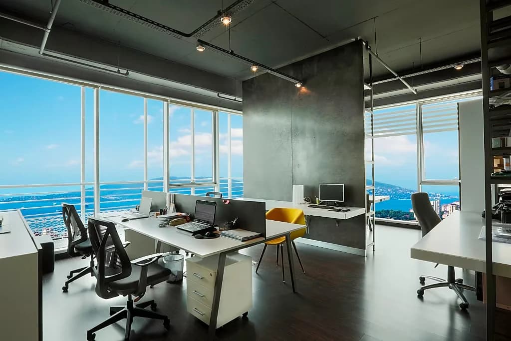 Open-plan office with desks along a glazed corner, a polished concrete column and a sea view beyond